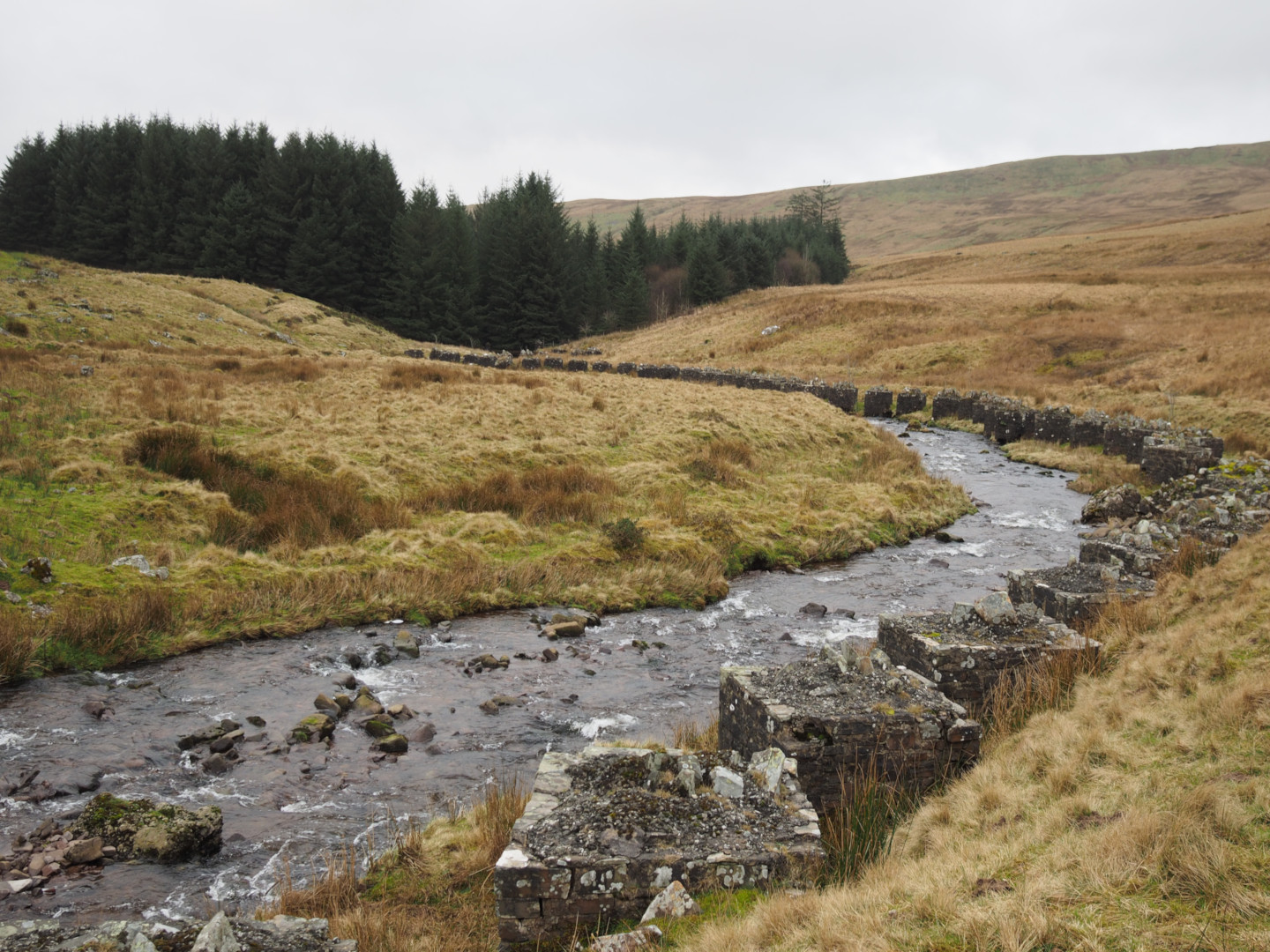 A photograph of a stream leading downhill, the stream lined with large cubes made of rock on one side. In the distance can be seen a large forested area, with hills in the background behind that.